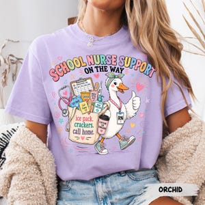 May include: A light purple t-shirt featuring a graphic of a white duck dressed as a school nurse. The duck wears a headband, an RN badge, and sneakers. It carries a tote bag filled with medical supplies, crackers, and a coffee cup with text.