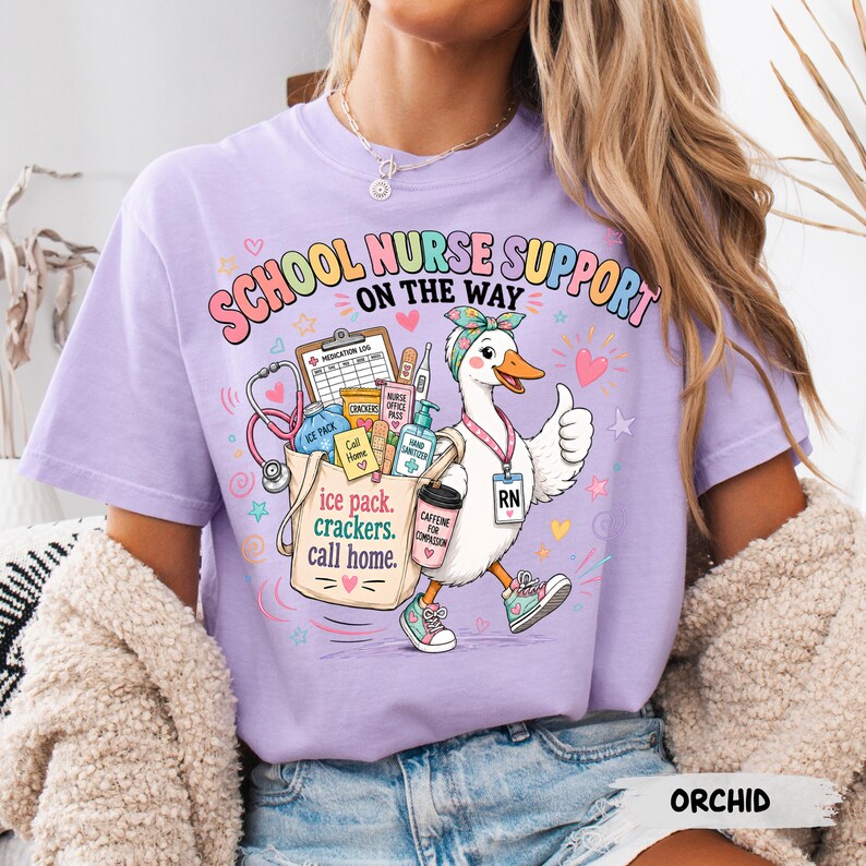 May include: A light purple t-shirt featuring a graphic of a white duck dressed as a school nurse. The duck wears a headband, an RN badge, and sneakers. It carries a tote bag filled with medical supplies, crackers, and a coffee cup with text.
