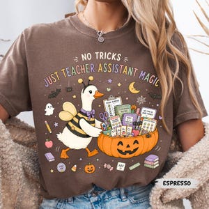 May include: A brown t-shirt featuring a graphic of a duck dressed as a bee standing next to a jack-o'-lantern filled with school supplies. The text reads NO TRICKS JUST TEACHER ASSISTANT MAGIC, surrounded by small icons of ghosts, bats, and books.
