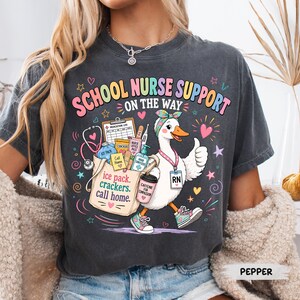 May include: A charcoal gray t-shirt featuring a graphic of a white duck wearing a headband, sneakers, and an RN badge. The duck carries a tote bag labeled 'ice pack, crackers, call home' with medical supplies. Text reads 'SCHOOL NURSE SUPPORT ON THE WAY'.