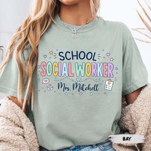K&ouml;nnte beinhalten: Ein salbeigr&uuml;nes Kurzarm-T-Shirt mit dem dunkelblauen Schriftzug SCHOOL und dem bunten Schriftzug SOCIAL WORKER. Darunter ist der Name Mrs. Mitchell in dunkelblauer Schreibschrift gedruckt, erg&auml;nzt durch kleine Symbole von Sternen, Herzen und einem Klemmbrett.
