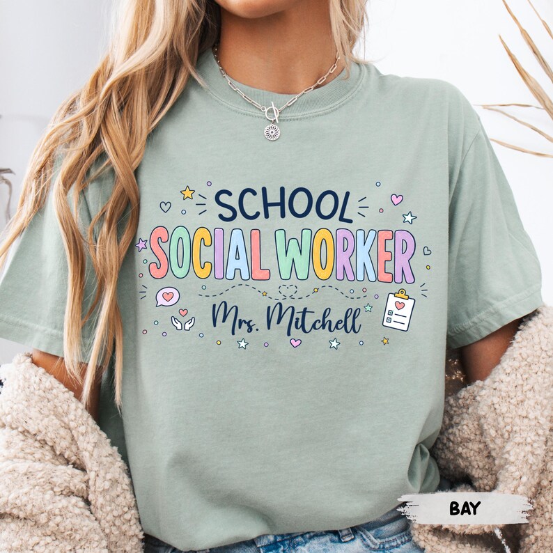 K&ouml;nnte beinhalten: Ein salbeigr&uuml;nes Kurzarm-T-Shirt mit dem dunkelblauen Schriftzug SCHOOL und dem bunten Schriftzug SOCIAL WORKER. Darunter ist der Name Mrs. Mitchell in dunkelblauer Schreibschrift gedruckt, erg&auml;nzt durch kleine Symbole von Sternen, Herzen und einem Klemmbrett.