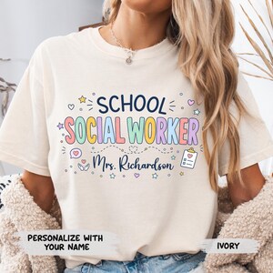 K&ouml;nnte beinhalten: Ein elfenbeinfarbenes Kurzarm-T-Shirt mit dem Text "SCHOOL SOCIAL WORKER" in bunten Buchstaben, "Mrs. Richardson" in Schreibschrift sowie kleinen Symbolen wie Sternen, Herzen, einer Sprechblase und einem Klemmbrett mit einem Herz.