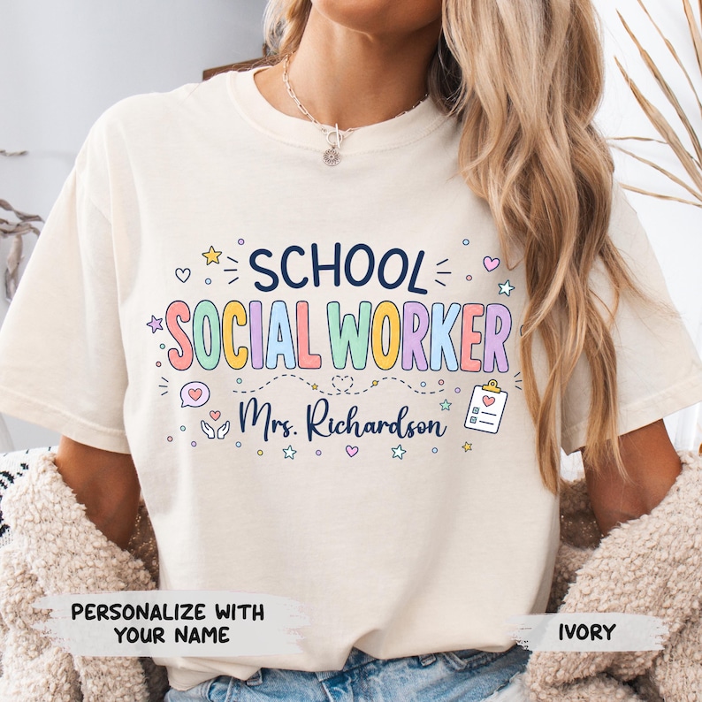 K&ouml;nnte beinhalten: Ein elfenbeinfarbenes Kurzarm-T-Shirt mit dem Text "SCHOOL SOCIAL WORKER" in bunten Buchstaben, "Mrs. Richardson" in Schreibschrift sowie kleinen Symbolen wie Sternen, Herzen, einer Sprechblase und einem Klemmbrett mit einem Herz.