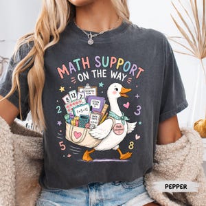 May include: Dark gray t-shirt with a colorful graphic of a goose carrying a bag of math supplies. The text "MATH SUPPORT ON THE WAY" is above the goose. Numbers and math symbols are scattered around the design. The word "PEPPER" is at the bottom.