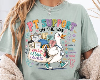 May include: A sage green t-shirt featuring a graphic of a white duck carrying a bag filled with physical therapy equipment. The text reads PT SUPPORT ON THE WAY, every step counts, KIDS FIRST, MOVE, STRENGTHEN, THRIVE, and SCHOOL PT.