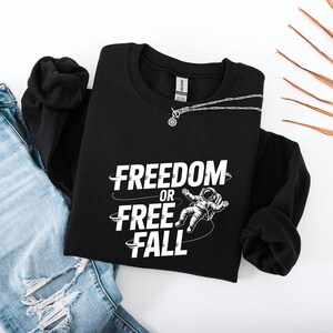 May include: Black crewneck sweatshirt with the text "FREEDOM OR FREE FALL" and an astronaut graphic in white. A silver necklace with a circular pendant is draped across the sweatshirt. The sweatshirt is paired with blue jeans.
