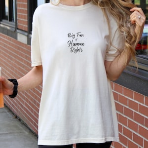 Pode incluir: Camiseta creme com o texto "Big Fan of Human Rights" em escrita preta. A camisa tem mangas curtas e um ajuste folgado. A pessoa está segurando um copo de plástico transparente com uma bebida marrom.