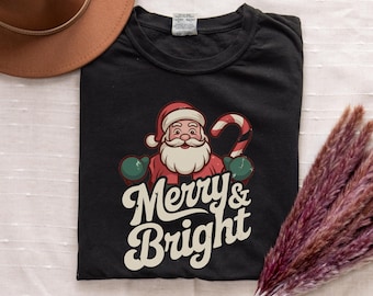 May include: Black t-shirt with a cartoon Santa Claus holding a candy cane and the text "Merry & Bright". The Santa graphic is red, white, and green. A brown hat and dried flowers are in the background.