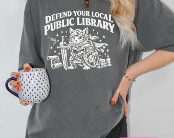 Puede incluir: Una camiseta gris carbón con un gráfico blanco de un gato vestido de caballero con armadura, sosteniendo una espada y un escudo con el icono de un edificio. El texto superior dice DEFEND YOUR LOCAL PUBLIC LIBRARY, junto a un pergamino y libros.