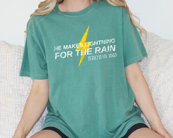 Puede incluir: Una camiseta de manga corta color verde azulado con un gráfico de un rayo amarillo centrado en el pecho. El texto blanco con efecto desgastado dice: HE MAKES LIGHTNING FOR THE RAIN JEREMIAH 10:13. La camiseta tiene cuello redondo y corte holgado.