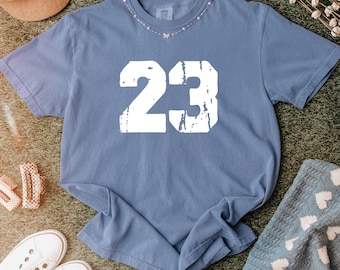 Puede incluir: Una camiseta azul de manga corta con el número 23 en letras grandes de bloque blanco con estilo desgastado, centradas en el pecho. La prenda tiene cuello redondo y un corte relajado, mostrando una estética de inspiración vintage con textura.