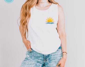 Puede incluir: Una camiseta sin mangas blanca con un pequeño gráfico en la zona del pecho izquierdo. El gráfico representa un sol amarillo saliendo sobre olas azules estilizadas. La camiseta se combina con unos vaqueros de mezclilla azul claro.
