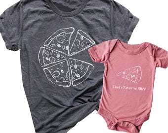 Puede incluir: Una camiseta gris oscuro con un gráfico de rebanada de pizza blanca y un body rosa para bebé con un gráfico de rebanada de pizza y el texto "Dad's Favorite Slice". Las camisetas están hechas de un material suave.