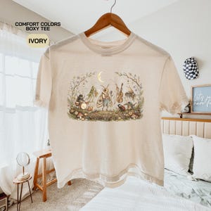 May include: An ivory boxy t-shirt featuring a graphic print of anthropomorphic insects playing musical instruments in a forest setting under a crescent moon. The text COMFORT COLORS BOXY TEE IVORY is printed in the upper left corner of the image.