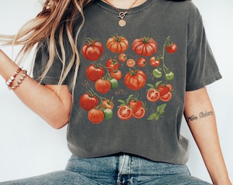 Puede incluir: Una camiseta de manga corta en color gris carbón con un estampado gráfico de varios tomates rojos en rama, incluyendo tomates enteros y cortados con tallos y hojas verdes, centrado en la parte delantera de la prenda.
