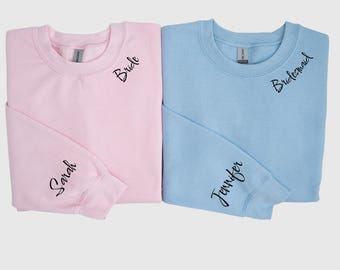 Puede incluir: Una sudadera de cuello redondo rosa claro con la palabra Bride impresa en el pecho y Sarah en la manga, junto a una sudadera azul claro con la palabra Bridesmaid en el pecho y Jennifer en la manga en letra cursiva negra.