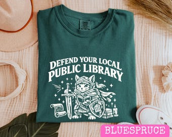 Puede incluir: Una camiseta verde oscuro con un gráfico blanco de un gato vestido de caballero con armadura, sosteniendo una espada y un libro, con un escudo que muestra un icono de edificio. El texto superior dice DEFEND YOUR LOCAL PUBLIC LIBRARY en blanco.