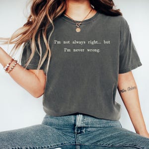 May include: Dark gray t-shirt with the text "I'm not always right... but I'm never wrong." The shirt is a crew neck style and appears to be made of a soft material. The model is wearing blue jeans and a gold necklace.