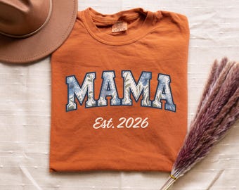 Puede incluir: Una camiseta de cuello redondo color naranja quemado con la palabra MAMA en un patrón floral azul y blanco con un contorno azul oscuro, y el texto blanco Est. 2026 debajo. Un sombrero de fieltro marrón y un manojo de hierba de la pampa seca.