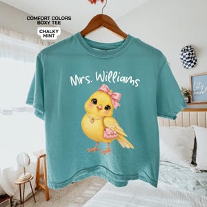 以下が含まれることがあります： チョークミント色のComfort ColorsボックスTシャツ。パールネックレスとピンクのリボンをつけ、ピンクのハンドバッグを持った黄色いヒヨコのグラフィックが描かれています。ヒヨコの上にはMrs. Williamsという白い文字がプリントされています。Tシャツはハンガーにかかっています。