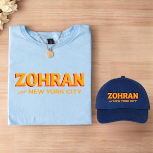 May include: A light blue t-shirt and a navy blue baseball cap. Both items feature the text "ZOHRAN FOR NEW YORK CITY" in orange and yellow. A silver necklace with a gold pendant is on the shirt.