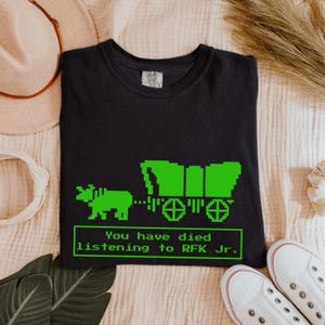 Puede incluir: Camiseta negra con una imagen pixelada verde de una carreta cubierta y bueyes. El texto "You have died listening to RFK Jr." se muestra en un cuadro verde. La camiseta es de color oscuro.