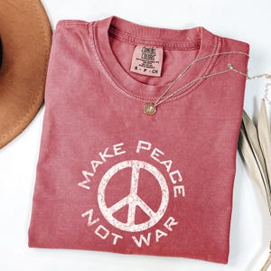May include: A dusty rose-colored t-shirt with the words "MAKE PEACE NOT WAR" printed in white, surrounding a peace symbol. A silver necklace with a small pendant rests on the shirt. The shirt is folded, and a brown hat is partially visible.