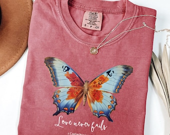 Puede incluir: Una camiseta de cuello redondo en color rosa empolvado con un gráfico grande y detallado de una mariposa con alas azules y naranjas. Debajo de la mariposa, el texto blanco dice "Love never fails" seguido de "1 Corinthians 13:8".