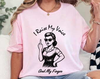 Puede incluir: Camiseta rosa claro con una ilustración en blanco y negro de una mujer con gafas de sol mostrando el dedo medio. El texto "I Raise My Voice And My Finger" está encima y debajo de la ilustración.