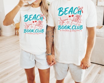 Puede incluir: Dos camisetas blancas de manga corta con un estampado gráfico con el texto BEACH BOOK CLUB sunshine & story time. El diseño incluye una silla de playa roja, un libro abierto, una pila de libros, gafas de sol, una bebida, una estrella de mar y un lazo rojo.