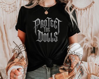 Puede incluir: Una camiseta negra de manga corta con el texto "PROTECT THE DOLLS" impreso en una fuente gótica gris metálico estilizada en el pecho. La camiseta tiene cuello redondo y el dobladillo está atado en un nudo en el centro delantero.