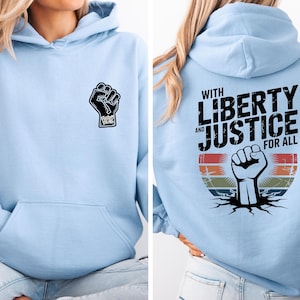 May include: Light blue hoodie with a black fist graphic on the front and back. The back of the hoodie features the text "WITH LIBERTY AND JUSTICE FOR ALL" above a fist graphic with a retro color scheme.