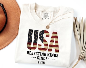 Puede incluir: Una camiseta de manga corta color blanco roto con el texto USA en un patrón de bandera estadounidense desgastado. Debajo de la bandera, el texto negro dice REJECTING KINGS SINCE 1776. La camiseta tiene una etiqueta Comfort Colors en el cuello.