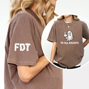 Puede incluir: Camiseta marrón con texto blanco y un gráfico de un fantasma haciendo una peineta. El texto de la camiseta dice "TO ALL RACISTS" y "FDT" en la manga. La camiseta es de estilo casual.