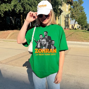 Könnte beinhalten: Ein grünes T-Shirt mit einer Grafik von mehreren Männern und dem Wort "ZOHRAN" in Gelb und Orange. Der Text "FOR NEW YORK CITY" steht unter dem Namen. Eine weiße Baseballkappe mit dem Wort "ZOHRAN" ist ebenfalls zu sehen.