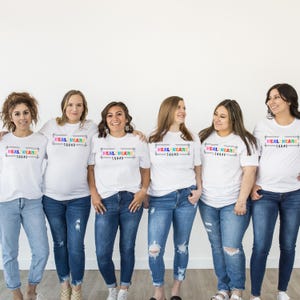Puede incluir: Seis personas con camisetas blancas y vaqueros azules. Cada camiseta tiene el texto "HEAL HCARE SQUAD" en un diseño rectangular colorido. Están de pie frente a una pared blanca.