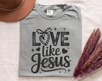 Puede incluir: Una camiseta gris de manga corta con el texto en negro LOVE like JESUS, un gráfico de lazo en forma de corazón, pequeñas estrellas y corazones adicionales. La camiseta tiene cuello redondo y una etiqueta de marca visible en el cuello.