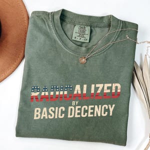 Op de afbeelding: Een olijfgroen t-shirt met de woorden "RADICALIZED" in rood en blauw, en "BY BASIC DECENCY" in wit. Een gouden ketting met een hanger ligt op het shirt. Het shirt is op een wit oppervlak gevouwen.