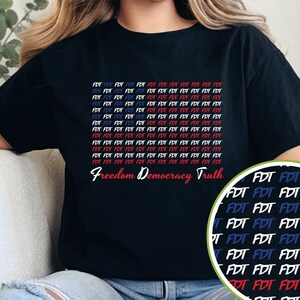Peut inclure: T-shirt noir avec les mots "Freedom Democracy Truth" en écriture blanche. Le t-shirt présente un motif répétitif de "FDT" en rouge, blanc et bleu, créant un design patriotique. Un gros plan du motif est dans le coin inférieur droit.