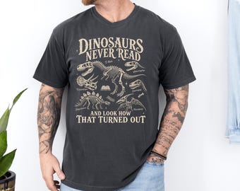 Puede incluir: Una camiseta gris carbón de manga corta con un gráfico color crema de varios esqueletos de dinosaurios. El texto dice: DINOSAURS NEVER READ AND LOOK HOW THAT TURNED OUT, con etiquetas para T-Rex, Triceratops, Velociraptor, Stegosaurus y Pterodactyl.