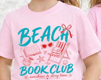 Puede incluir: Una camiseta rosa claro con un gráfico que incluye el texto turquesa BEACH BOOK CLUB y el texto rojo sunshine & story time. El diseño incluye un libro abierto rojo, gafas de sol rojas, una silla de playa roja con libros, una estrella de mar.