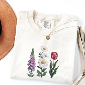May include: Cream-colored Comfort Colors t-shirt featuring a floral design with a foxglove, daisies, and a tulip. A gold necklace with a pendant rests on the shirt. The shirt tag reads "Comfort Colors".