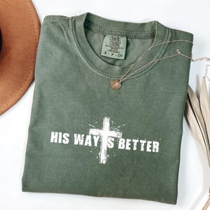 May include: An olive green t-shirt with the text "HIS WAY'S BETTER" in white, featuring a white cross graphic. A gold necklace with a pendant rests on the shirt. A brown hat is visible in the upper left corner.