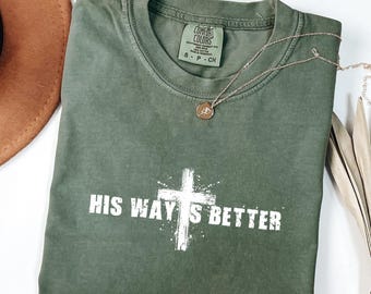 May include: An olive green t-shirt with the text "HIS WAY'S BETTER" in white, featuring a white cross graphic. A gold necklace with a pendant rests on the shirt. A brown hat is visible in the upper left corner.