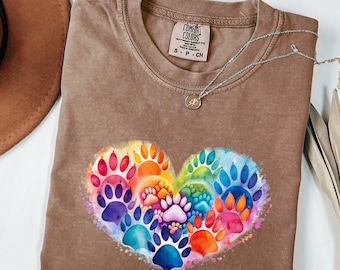 Puede incluir: Una camiseta Comfort Colors marrón con un diseño de corazón compuesto por huellas de patas multicolores. Las huellas de patas son en tonos de rojo, naranja, amarillo, verde, azul y morado. Un collar plateado con un pequeño colgante está sobre la camisa.