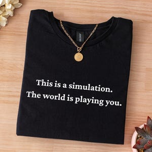 Puede incluir: Camiseta negra con el texto "This is a simulation. The world is playing you." Un collar dorado con un colgante redondo descansa sobre la camiseta. La camiseta está doblada sobre una superficie de madera.