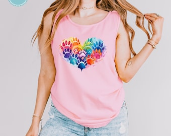 Puede incluir: Una camiseta sin mangas de color rosa claro con un gráfico central en forma de corazón compuesto por huellas de animales estilo acuarela en tonos azules, verdes, amarillos, naranjas y rojos. La prenda se combina con unos vaqueros azul claro.