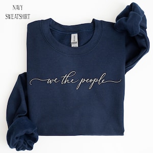 May include: A navy blue crewneck sweatshirt featuring the text "we the people" in a white cursive script font across the center chest. The garment has long sleeves and a ribbed neckline, with a Gildan brand label visible at the inner collar.