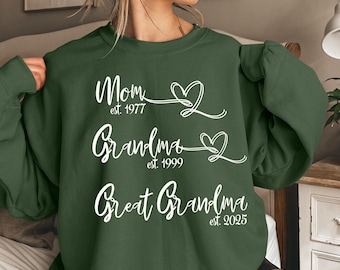 Puede incluir: Una sudadera de manga larga color verde bosque con texto blanco y gráficos de corazones. El estampado muestra las palabras Mom est. 1977, Grandma est. 1999 y Great Grandma est. 2025 en una fuente cursiva, cada una con un contorno de corazón.